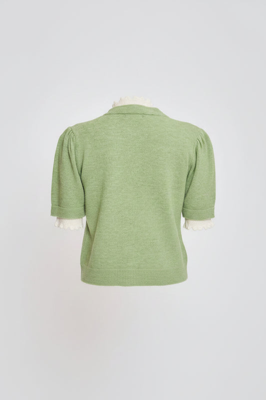 A woman shown from the back wearing a light green short-sleeve knit top with white ruffled trim at the collar and cuffs, paired with a cream pleated skirt, against a light gray studio background.