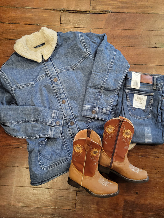 A flat-lay arrangement of clothing and footwear on a wooden floor: a blue denim jacket with a cream sherpa collar, a pair of blue jeans, and a pair of tan western cowboy boots with embroidered sun motifs.