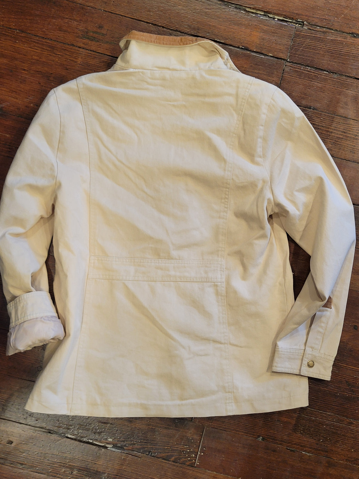 A cream canvas Barn jacket/coat with a high collar and long sleeves, Back Side, laid flat on a rustic dark wood floor.