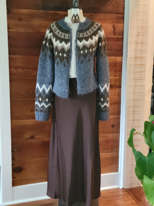 A blue-gray knit cardigan with a brown, white, and gray Fair Isle pattern, styled over a white top and a dark brown skirt on a dress form, set against a wooden paneled wall.