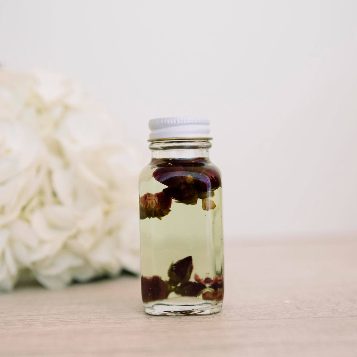 Glass bath oil bottle containing dark red petals beside white flowers