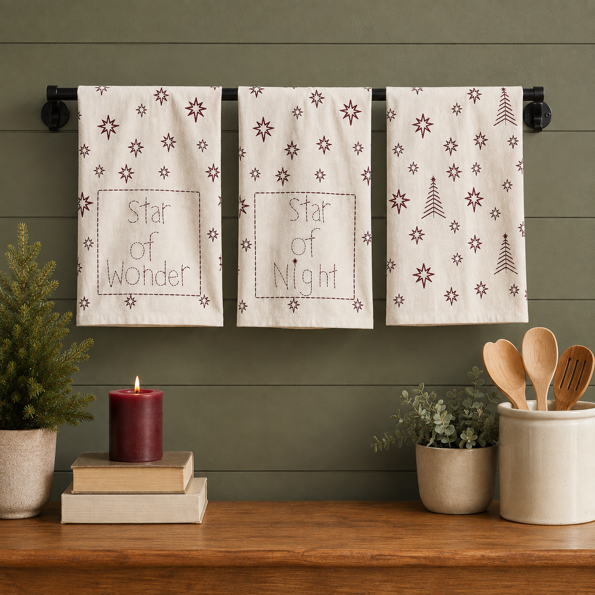 Three cream Christmas tea towel styles with burgundy stars and trees hanging above a holiday kitchen counter.