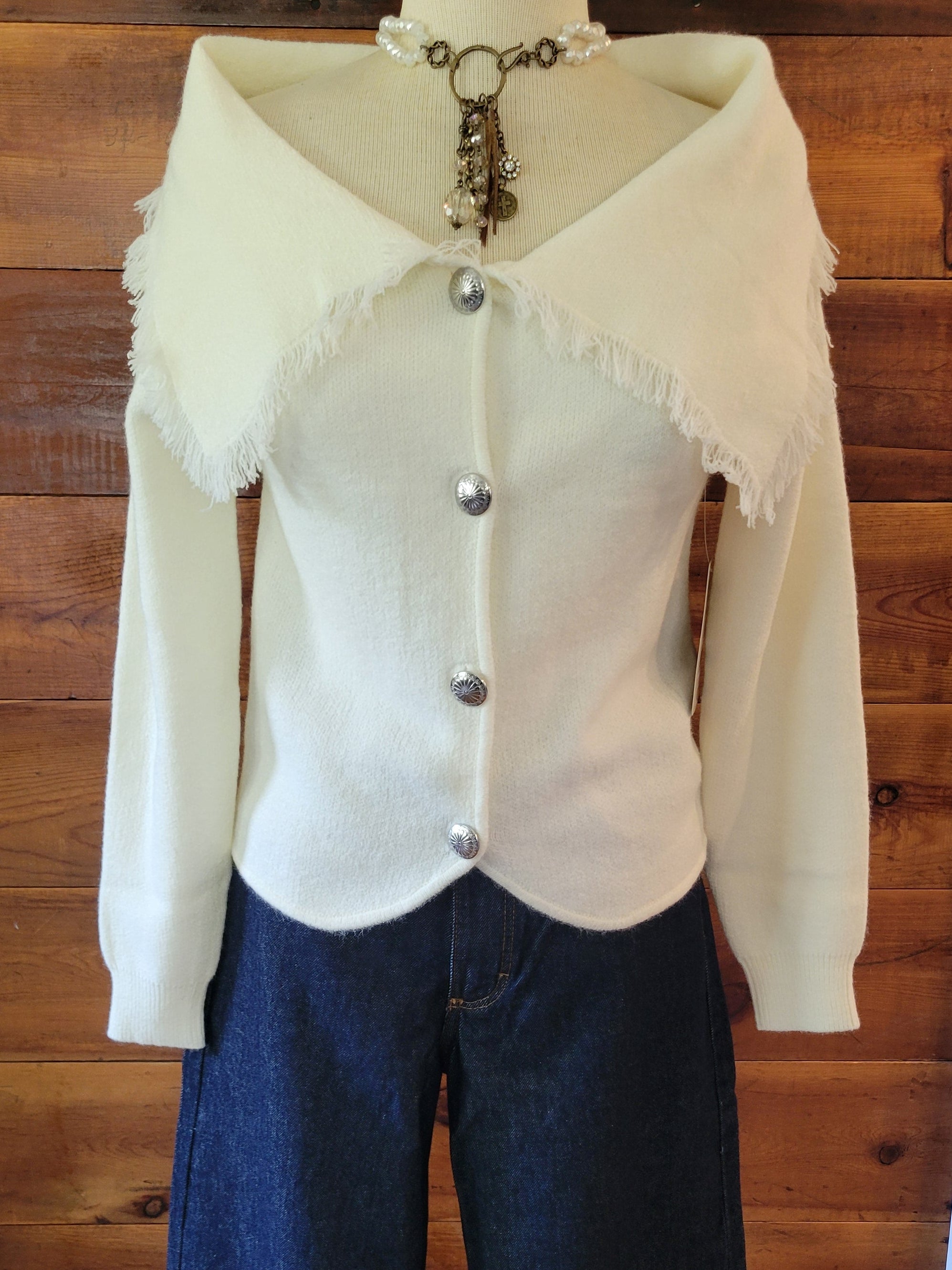 A cream knit cardigan with a wide frayed collar and silver buttons, styled with dark blue wide-leg jeans on a mannequin in front of a wooden wall.