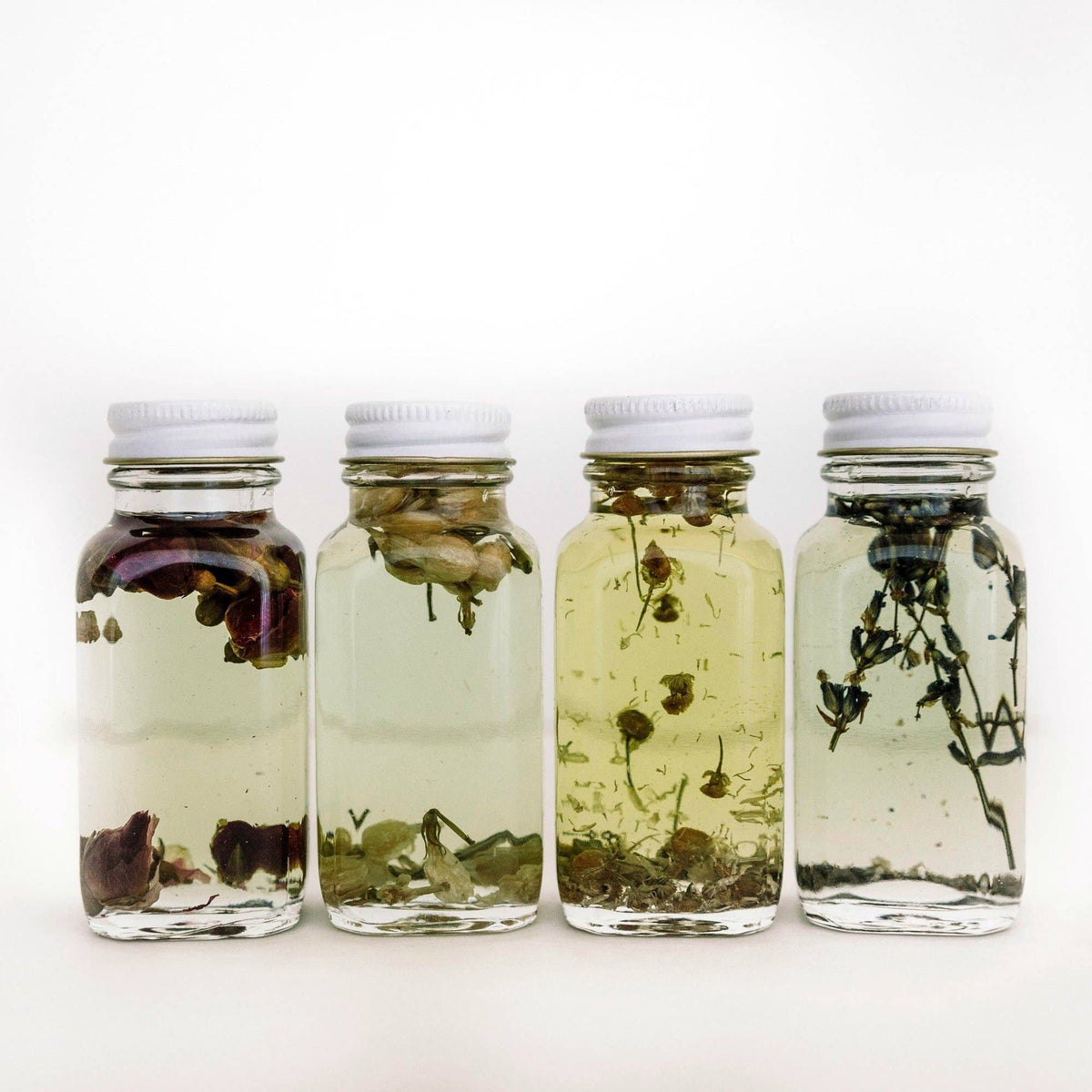 Four glass bath oil bottles with botanical pieces and white caps on a white background