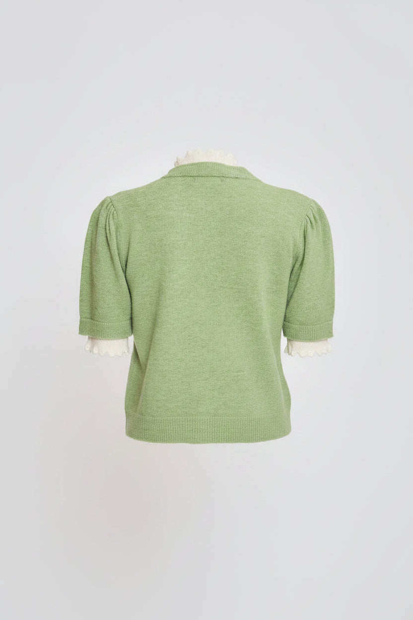 A woman shown from the back wearing a light green short-sleeve knit top with white ruffled trim at the collar and cuffs, paired with a cream pleated skirt, against a light gray studio background.