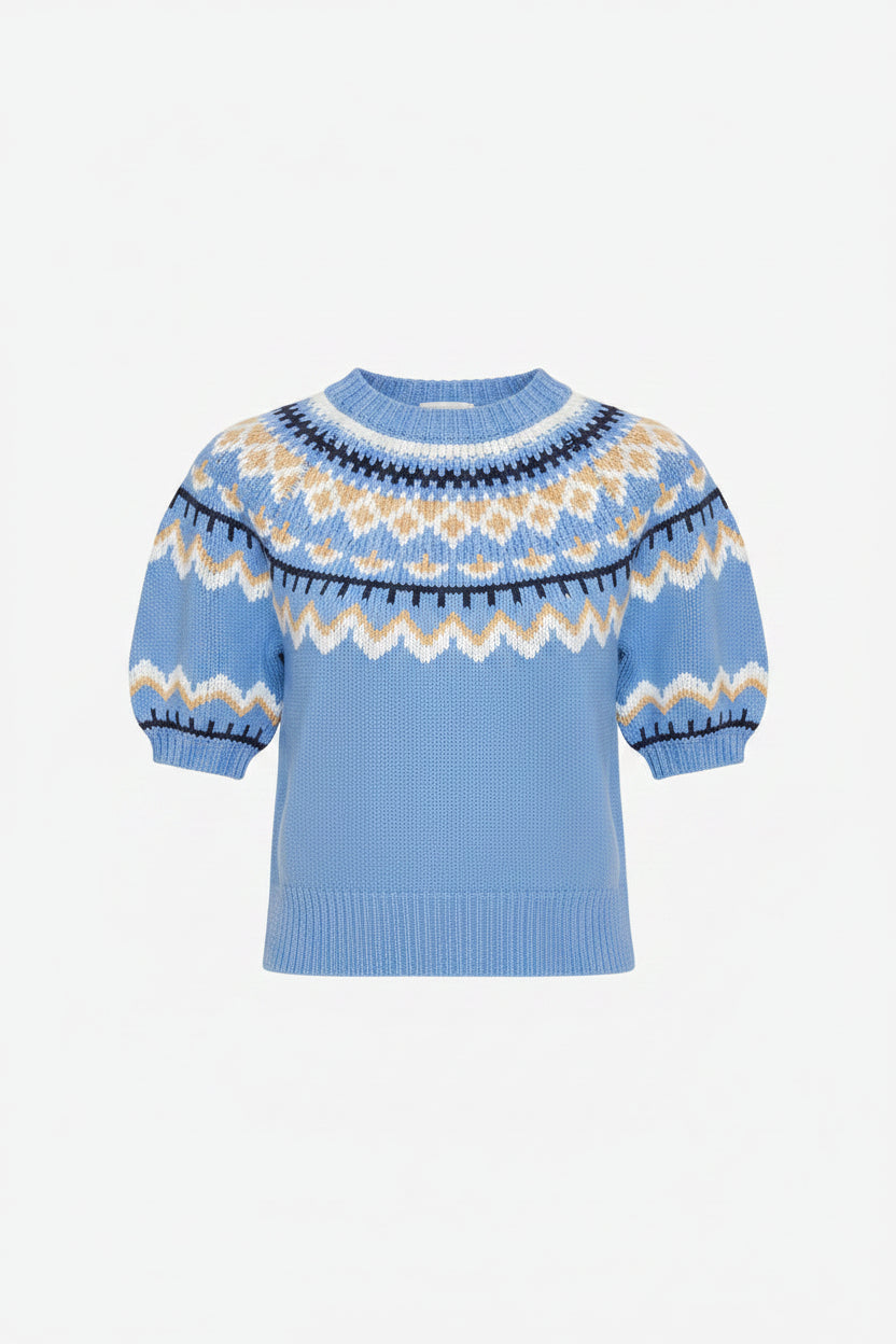 A woman wearing a light blue short-sleeve knit sweater with a white and yellow geometric pattern around the collar and cuffs, paired with a white tiered skirt, against a light gray studio background.