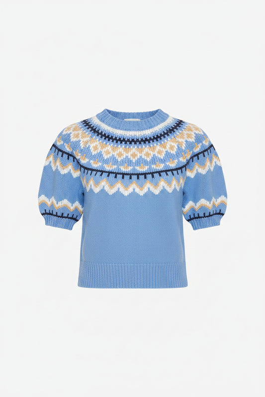 A woman wearing a light blue short-sleeve knit sweater with a white and yellow geometric pattern around the collar and cuffs, paired with a white tiered skirt, against a light gray studio background.
