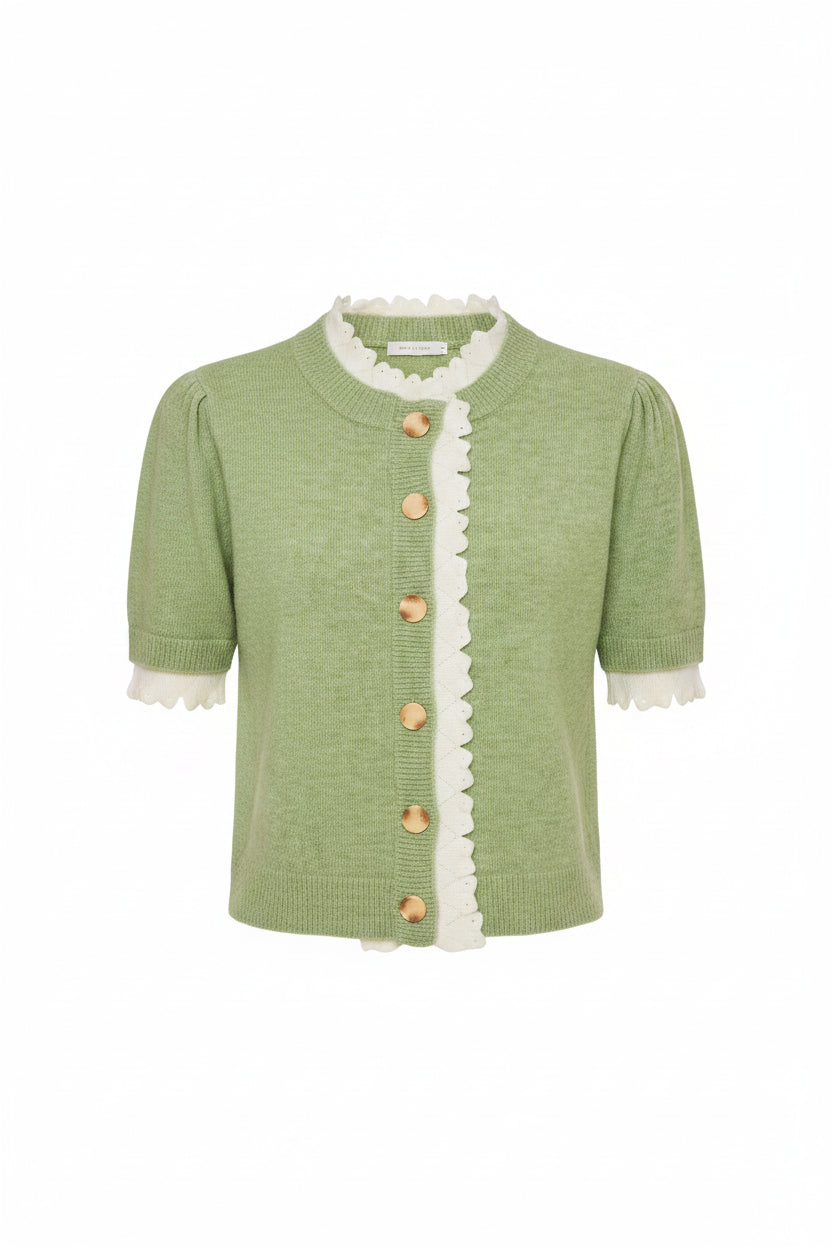A woman modeling a light green short-sleeve knit cardigan with white scalloped trim and wooden buttons, paired with cream shorts, against a light gray studio background.