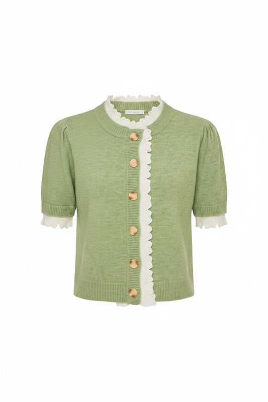 A woman modeling a light green short-sleeve knit cardigan with white scalloped trim and wooden buttons, paired with cream shorts, against a light gray studio background.