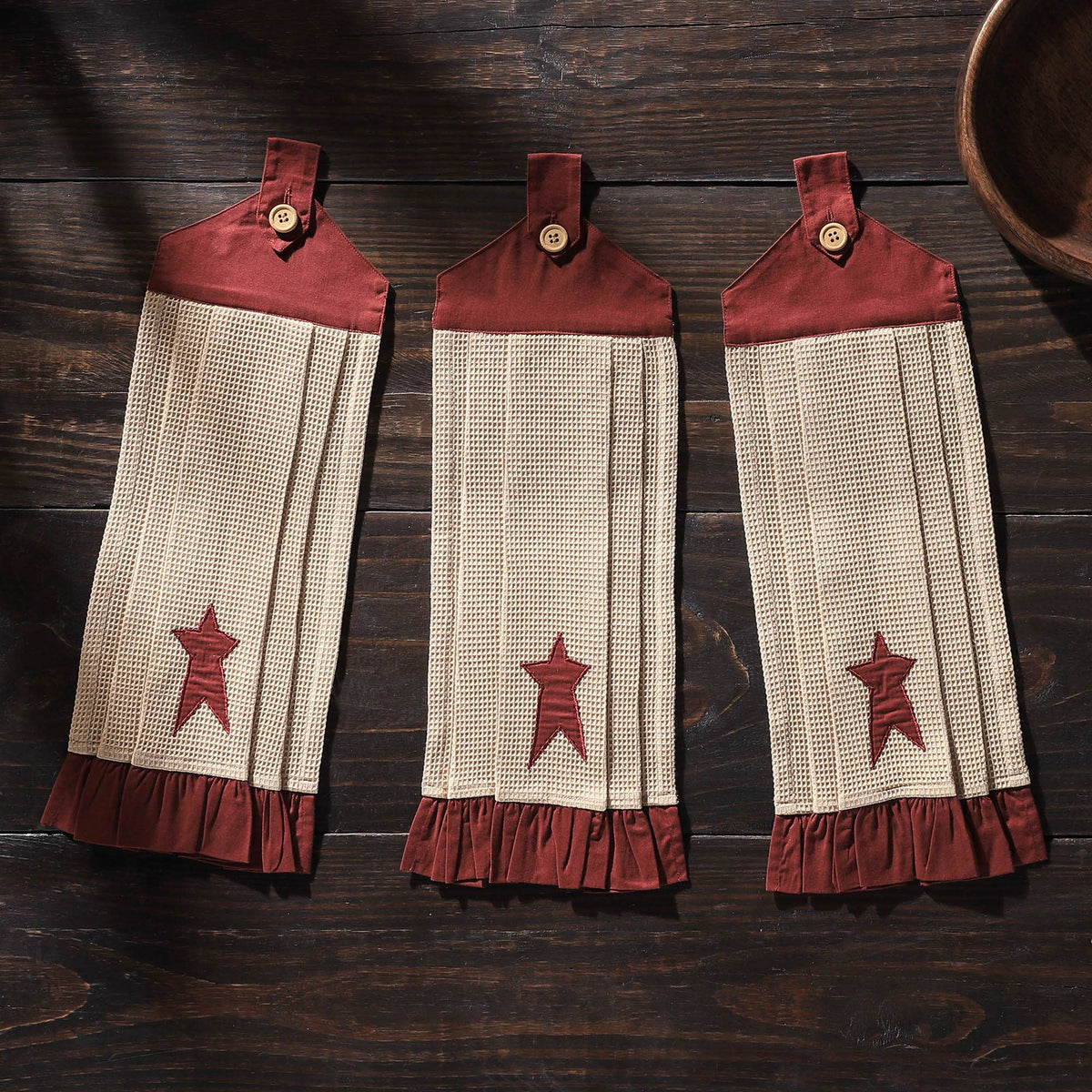 Three Connell cream waffle-textured kitchen towels with burgundy stars, button loops, and ruffled hems displayed on dark wood.