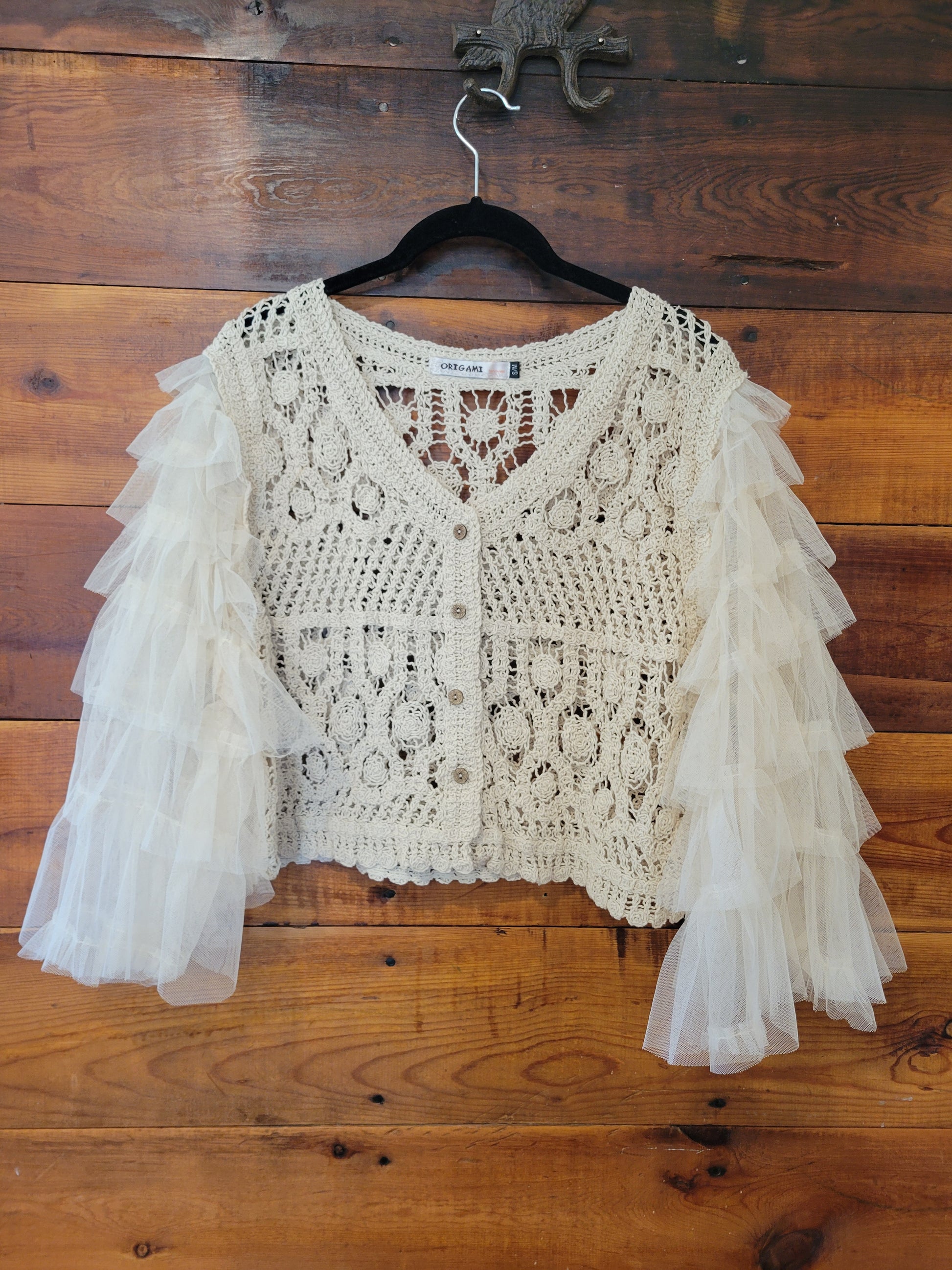 An off-white crochet cardigan sweater with layered tulle ruffle sleeves, laid flat on a black hanger over a rustic wooden plank floor.