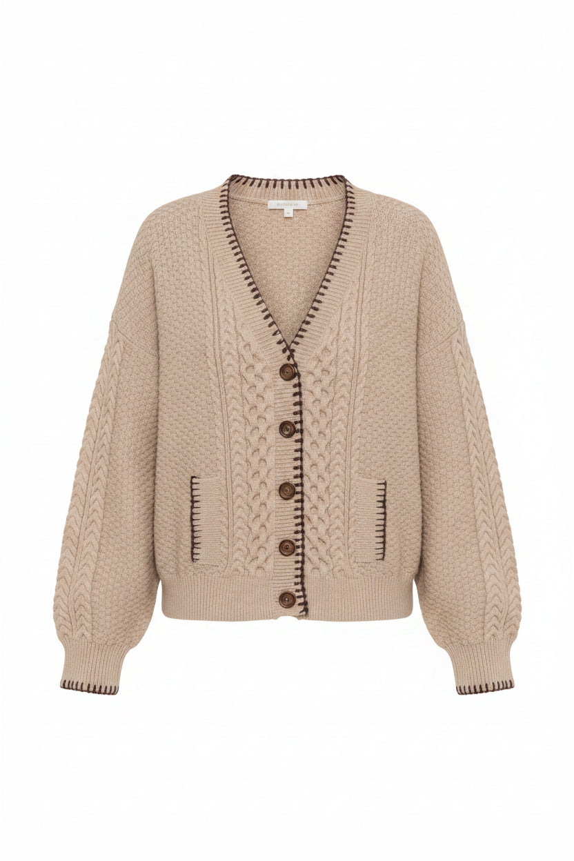 A woman wearing a beige cable-knit cardigan sweater with dark brown buttons and trim, paired with brown pants, against a plain white studio background.