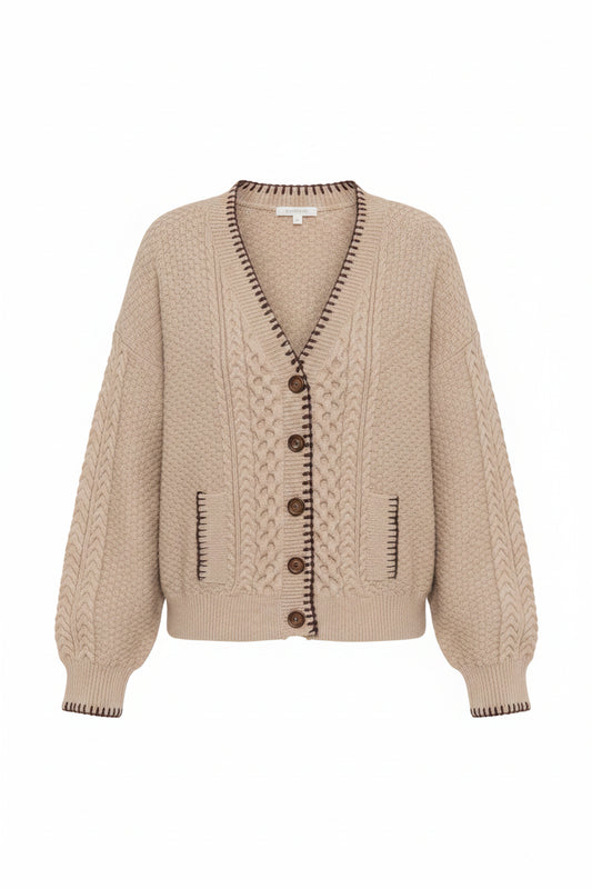 A woman wearing a beige cable-knit cardigan sweater with dark brown buttons and trim, paired with brown pants, against a plain white studio background.