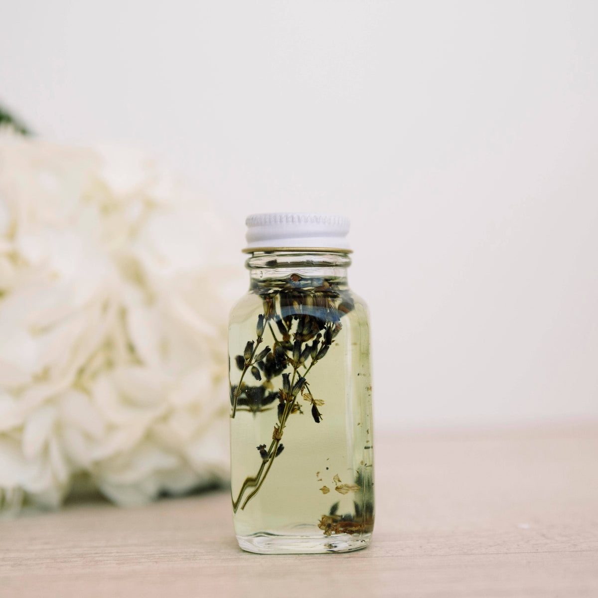 Glass bath oil bottle containing dark botanical stems beside white flowers