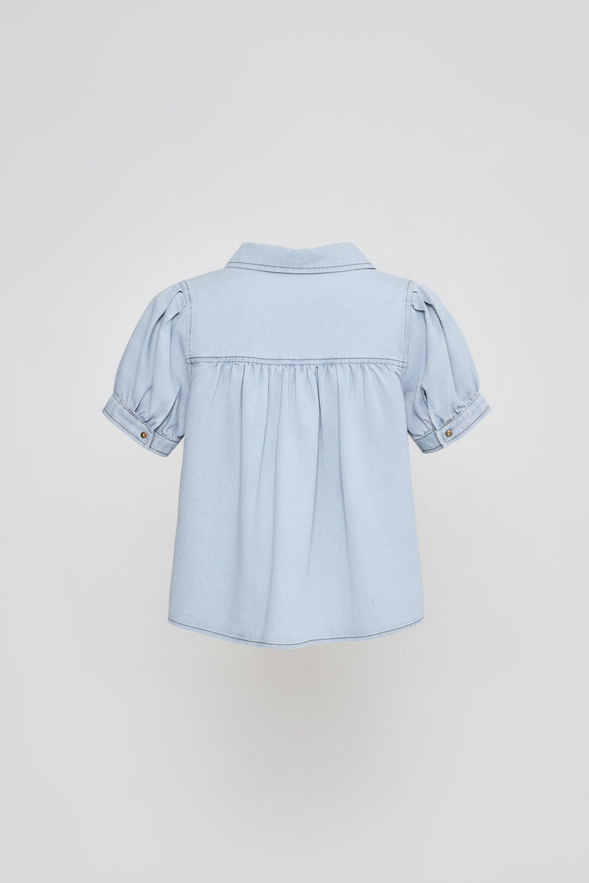 A woman shown from the back wearing a light blue cropped short-sleeve blouse with puffed sleeves and white wide-leg pants, against a plain light gray studio background.