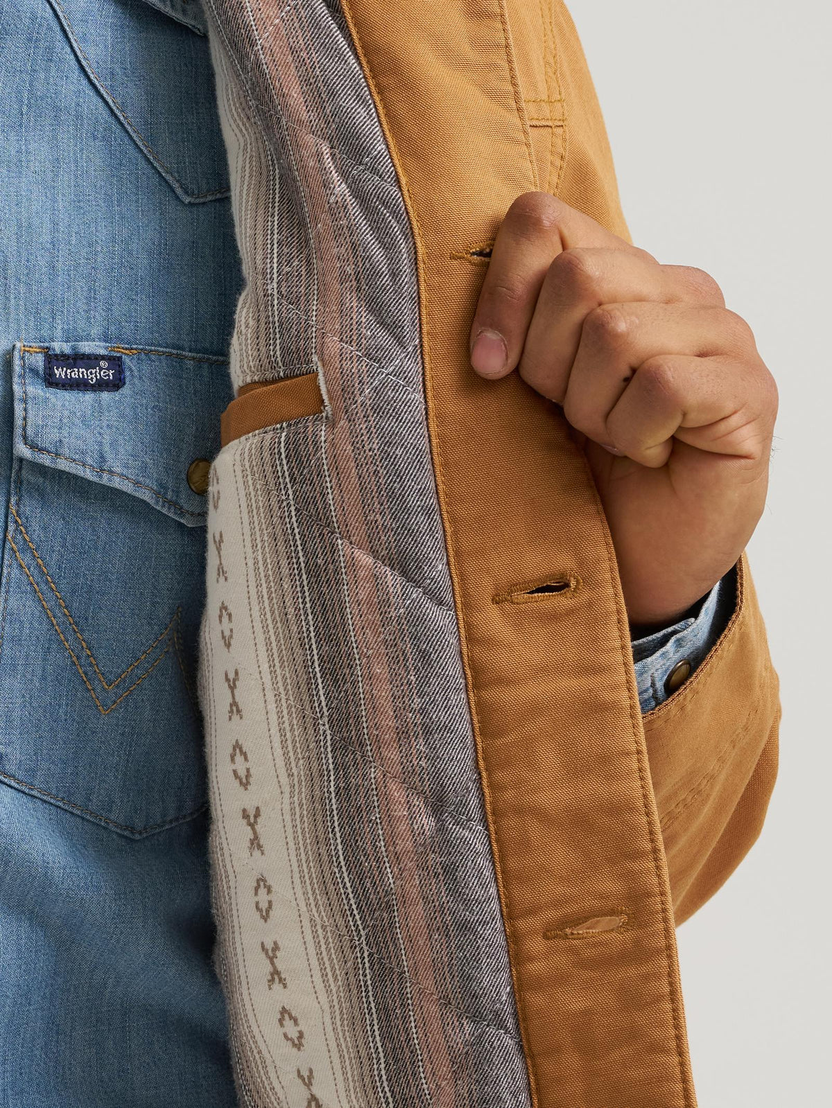 Close-up of a person wearing a tan jacket over a blue denim shirt, with the jacket's striped quilted lining and a hand holding the edge open.