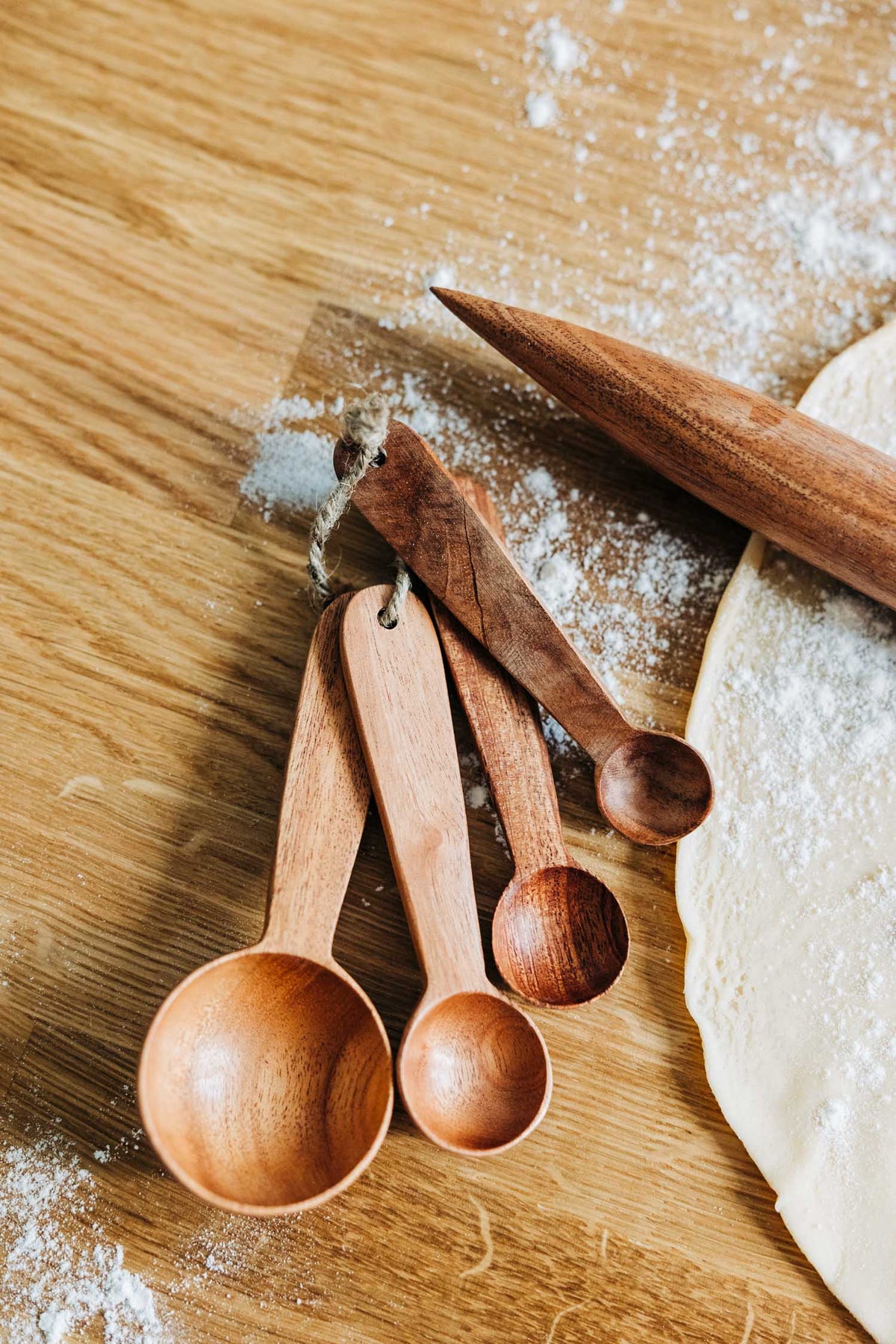 Four neem wood measuring spoons beside a rolling pin and dough on a flour-dusted wooden surface