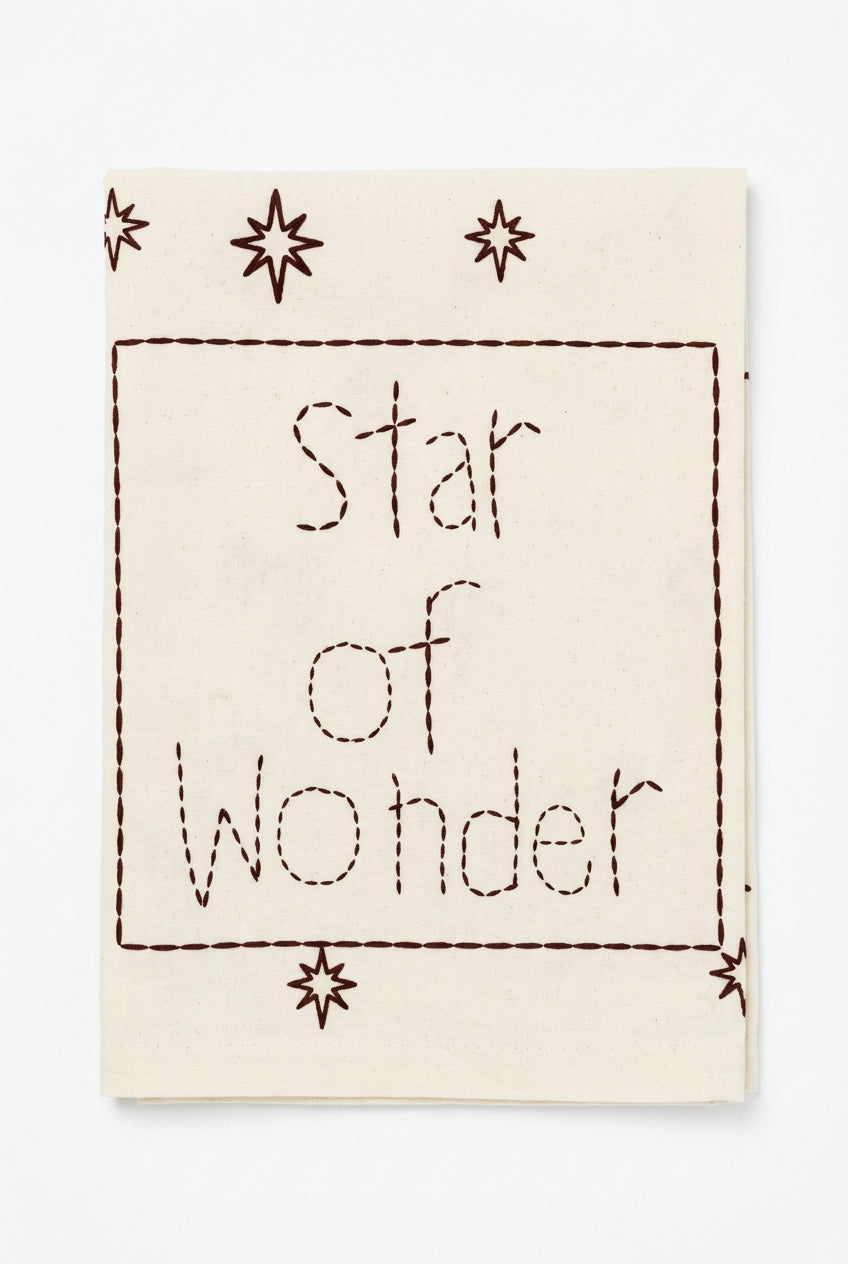 Folded cream Star of Wonder Christmas tea towel with burgundy lettering, star motifs, and a stitch-style printed border