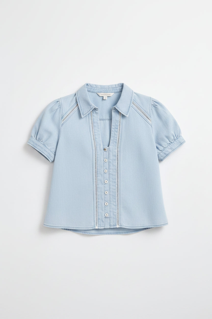 A woman modeling a light blue short-sleeve button-front blouse and white cropped pants with floral embroidery and lace trim, paired with beige flats, against a light gray studio background.