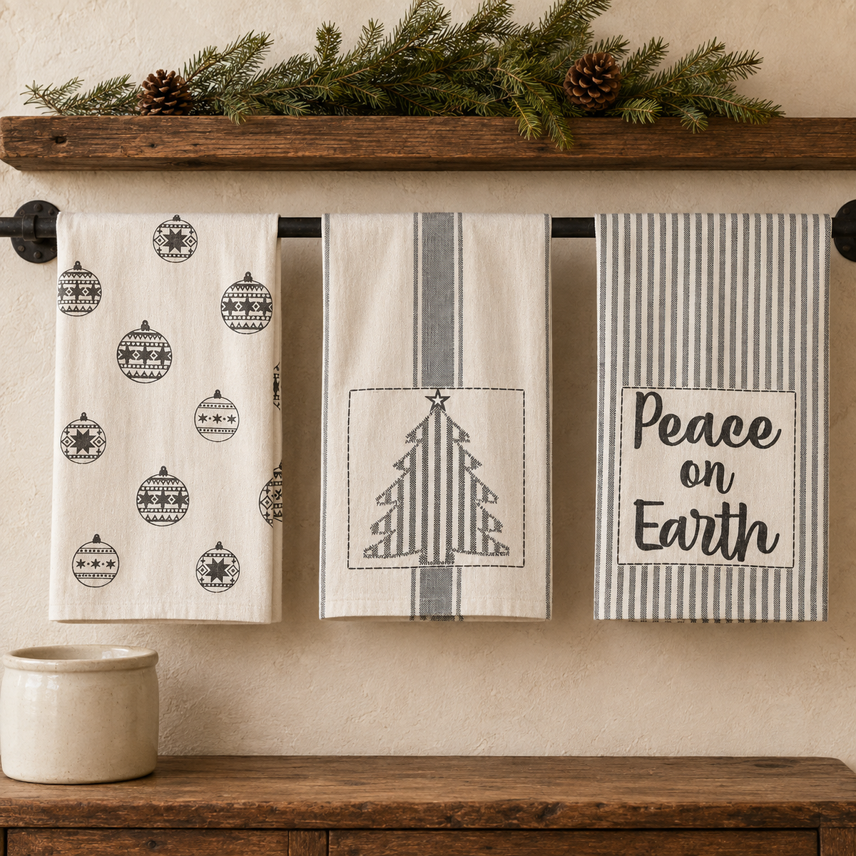 Three Grace Christmas tea towel styles hanging beneath greenery, featuring gray ornaments, a striped tree, and Peace on Earth lettering.