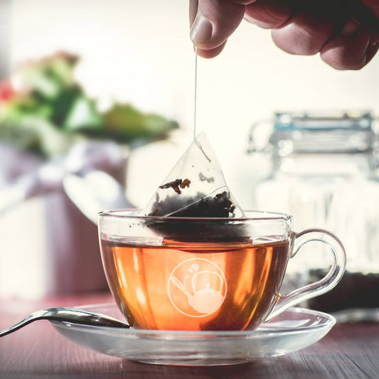 Hand lifting a pyramid tea sachet from amber tea in a clear glass cup and saucer