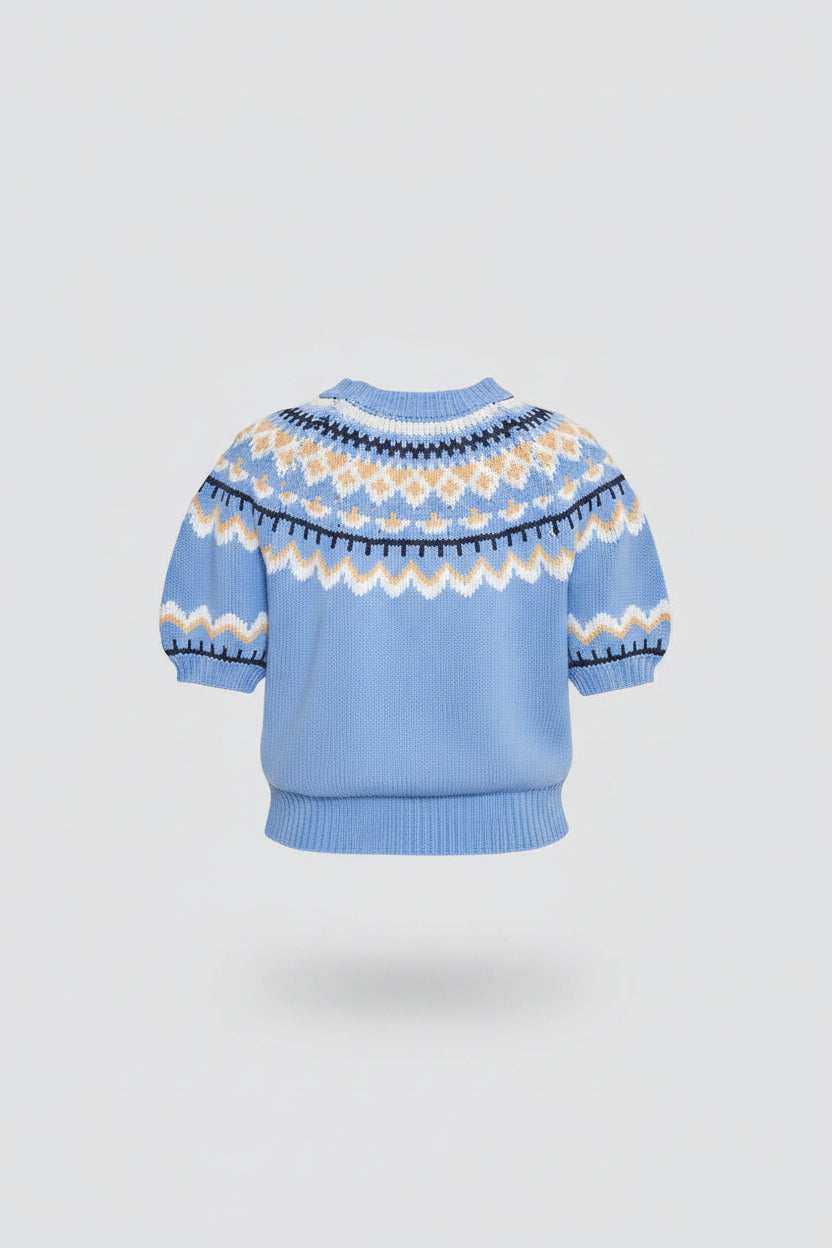A woman shown from the back wearing a light blue short-sleeve knit sweater with a cream and navy Fair Isle pattern across the shoulders and upper back, paired with a white tiered skirt, against a light gray studio background.