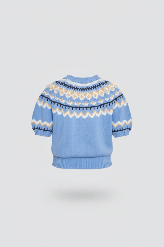 A woman shown from the back wearing a light blue short-sleeve knit sweater with a cream and navy Fair Isle pattern across the shoulders and upper back, paired with a white tiered skirt, against a light gray studio background.