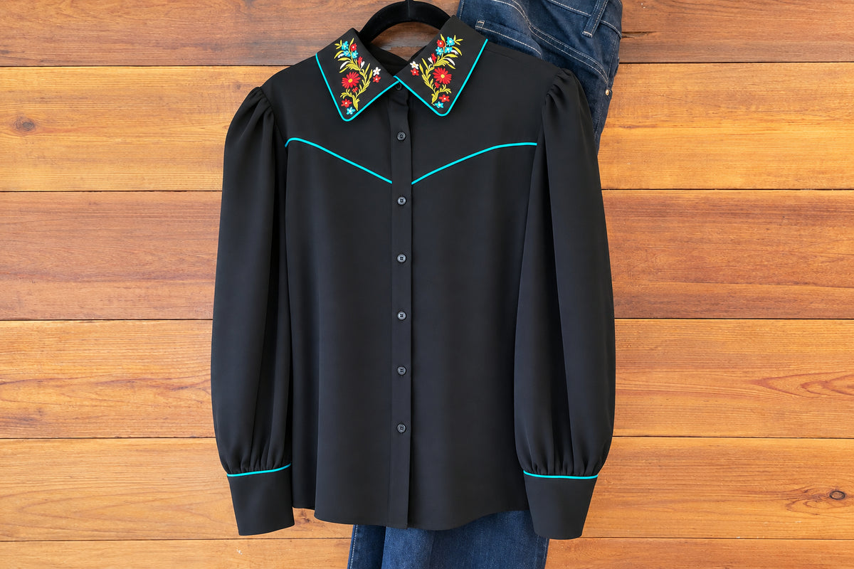 A black western-style blouse with turquoise piping and a colorful floral embroidered collar, shown hanging against a wooden plank wall. A denim skirt and jeans are partially visible underneath.