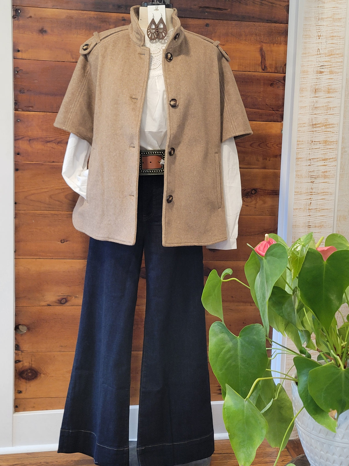 A styled outfit displayed on a mannequin or hanger against a wood-paneled wall, featuring a Toffee short-sleeve button-front cape jacket over a white blouse, dark navy wide-leg pants, and a brown belt. A green potted plant is visible in the foreground on the right.