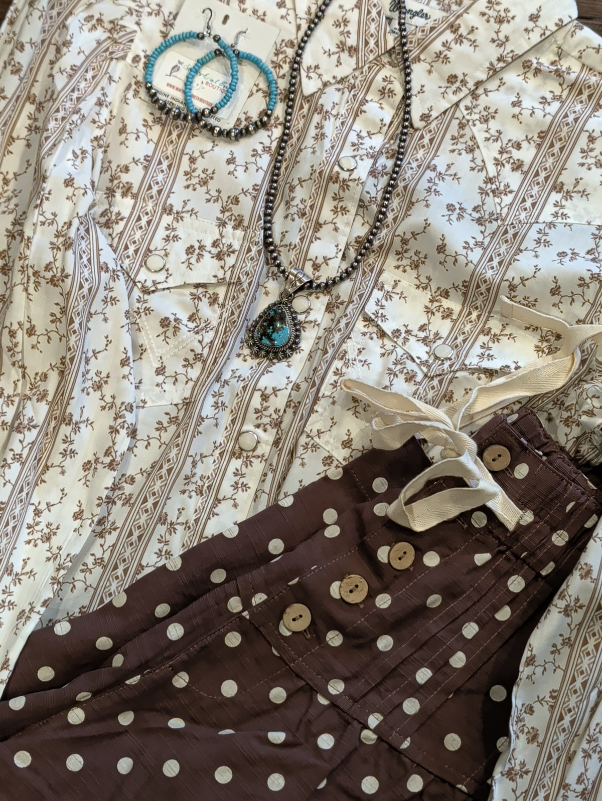 Close-up of brown polka dot pants showing front buttons and a cream waist tie beside a floral blouse and jewelry.