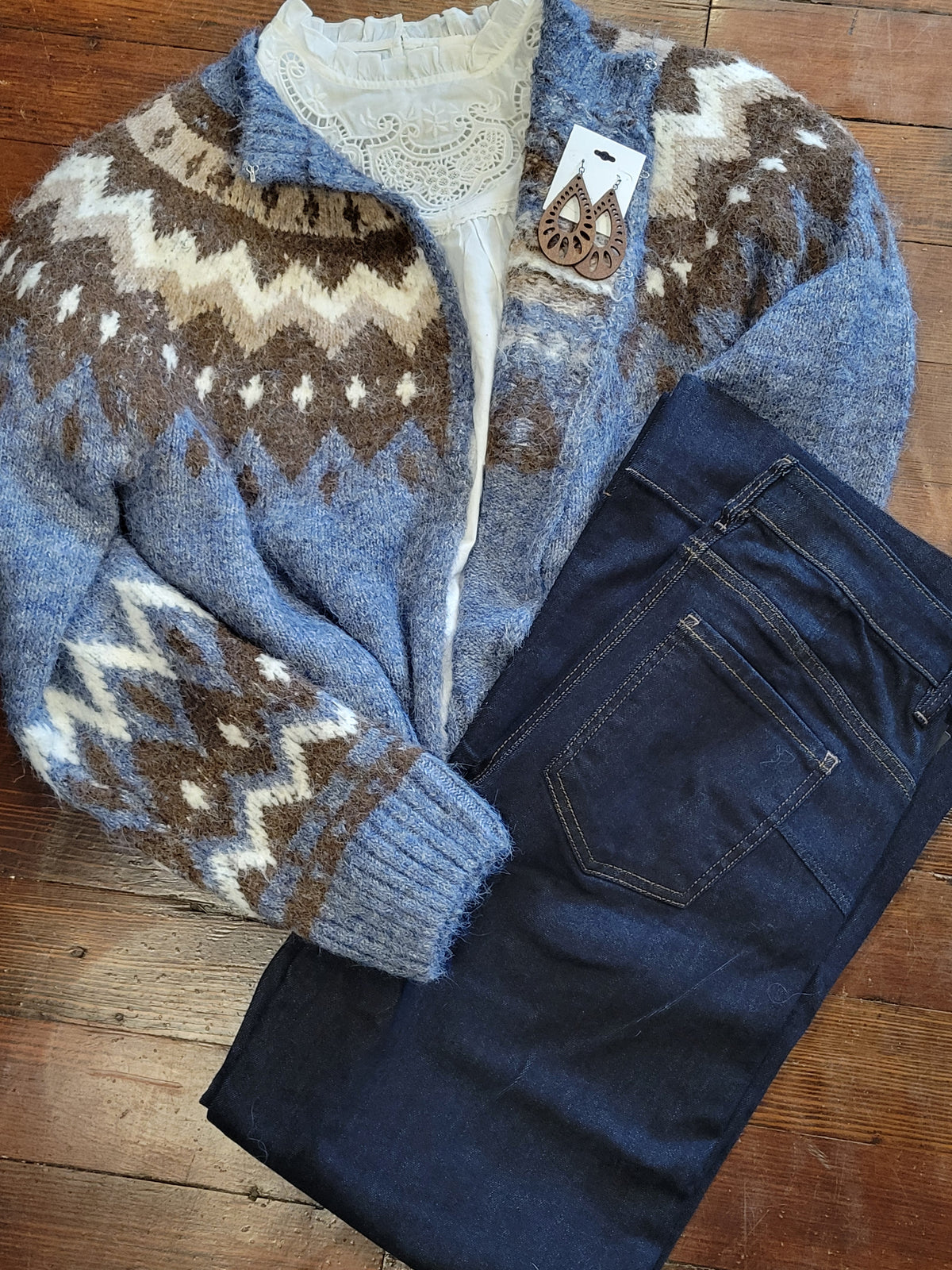 A folded knit cardigan with a blue, brown, and cream Fair Isle pattern is laid flat on a wooden floor next to a pair of dark blue jeans. A small wooden brand tag with wooden earrings is placed near the collar.