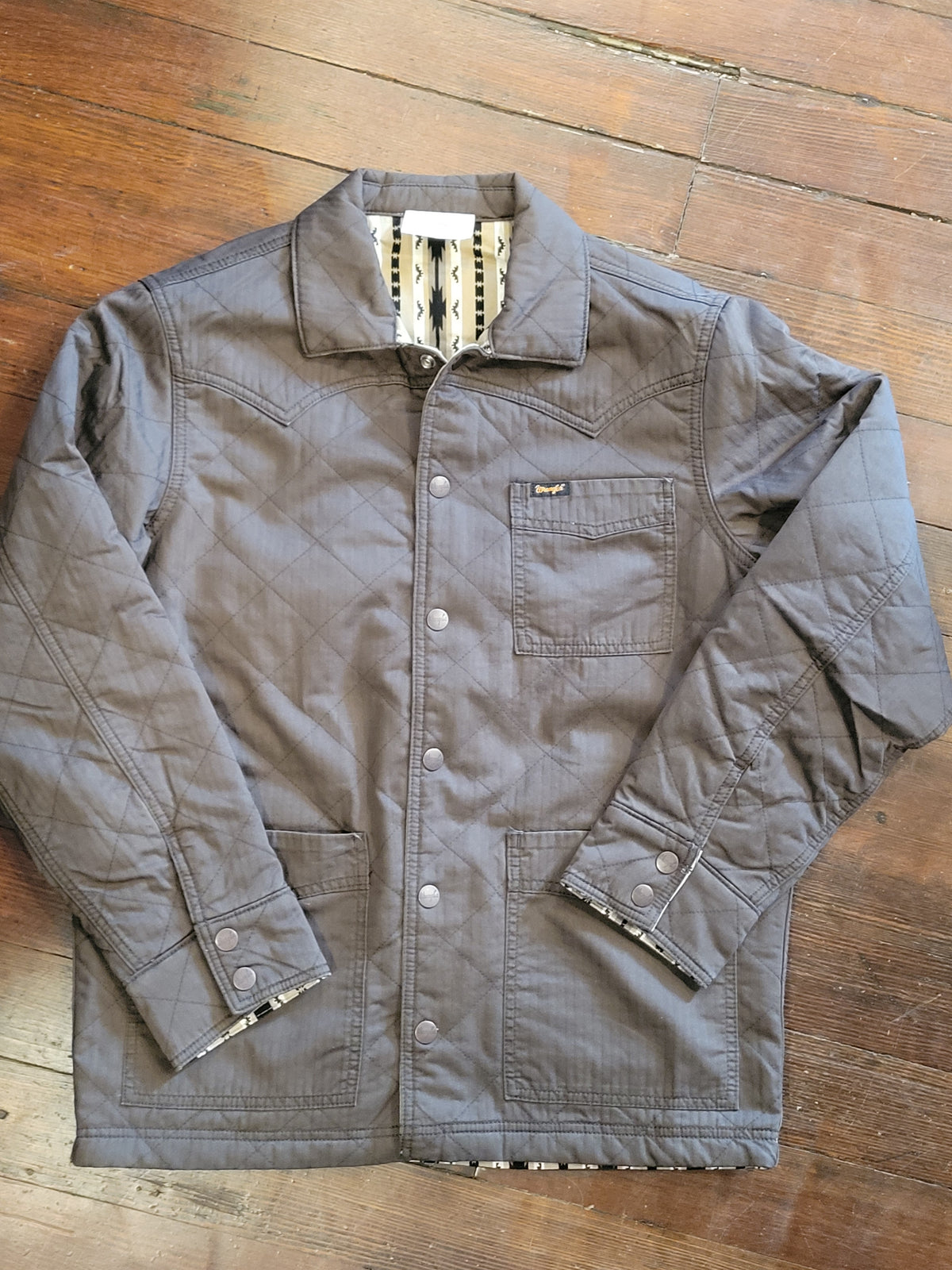 A dark brown quilted overshirt jacket with a button-up front, chest pocket, and patterned lining, laid flat on a wooden floor.