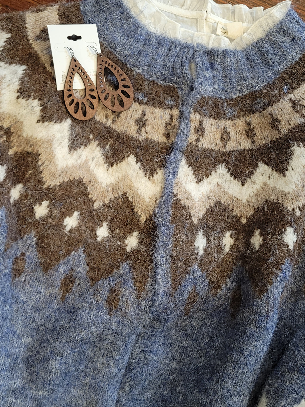 A close-up of a fuzzy knit cardigan sweater with a blue, brown, and cream Fair Isle pattern, featuring a white paper tag with brown teardrop-shaped wooden earrings attached.