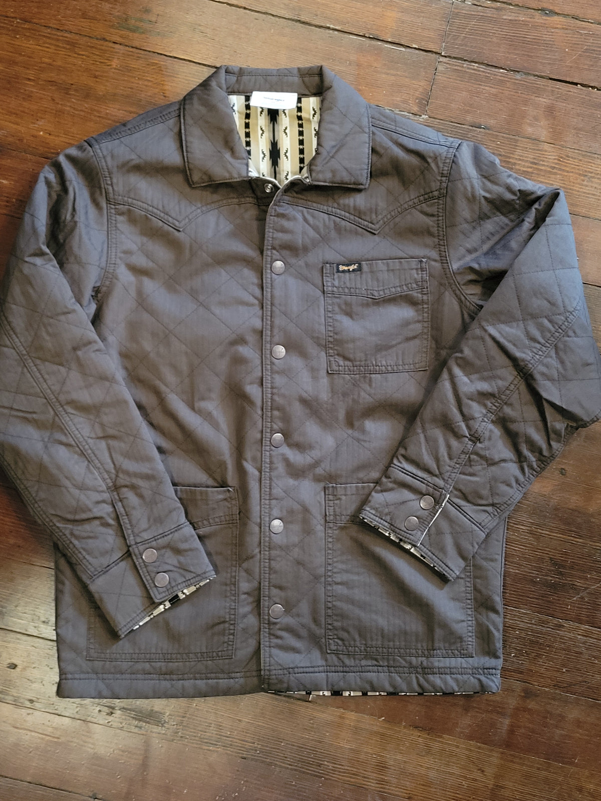 A dark brown quilted jacket with a button-up front and chest pocket, laid flat on a wooden floor.