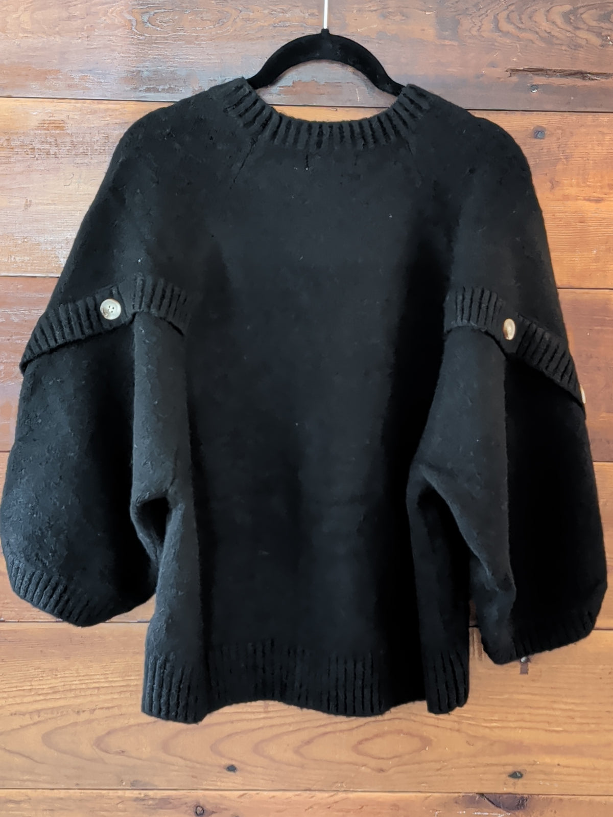 Black knit pullover on a hanger with wide sleeves folded forward to show ribbed trim and button details showing the back view.