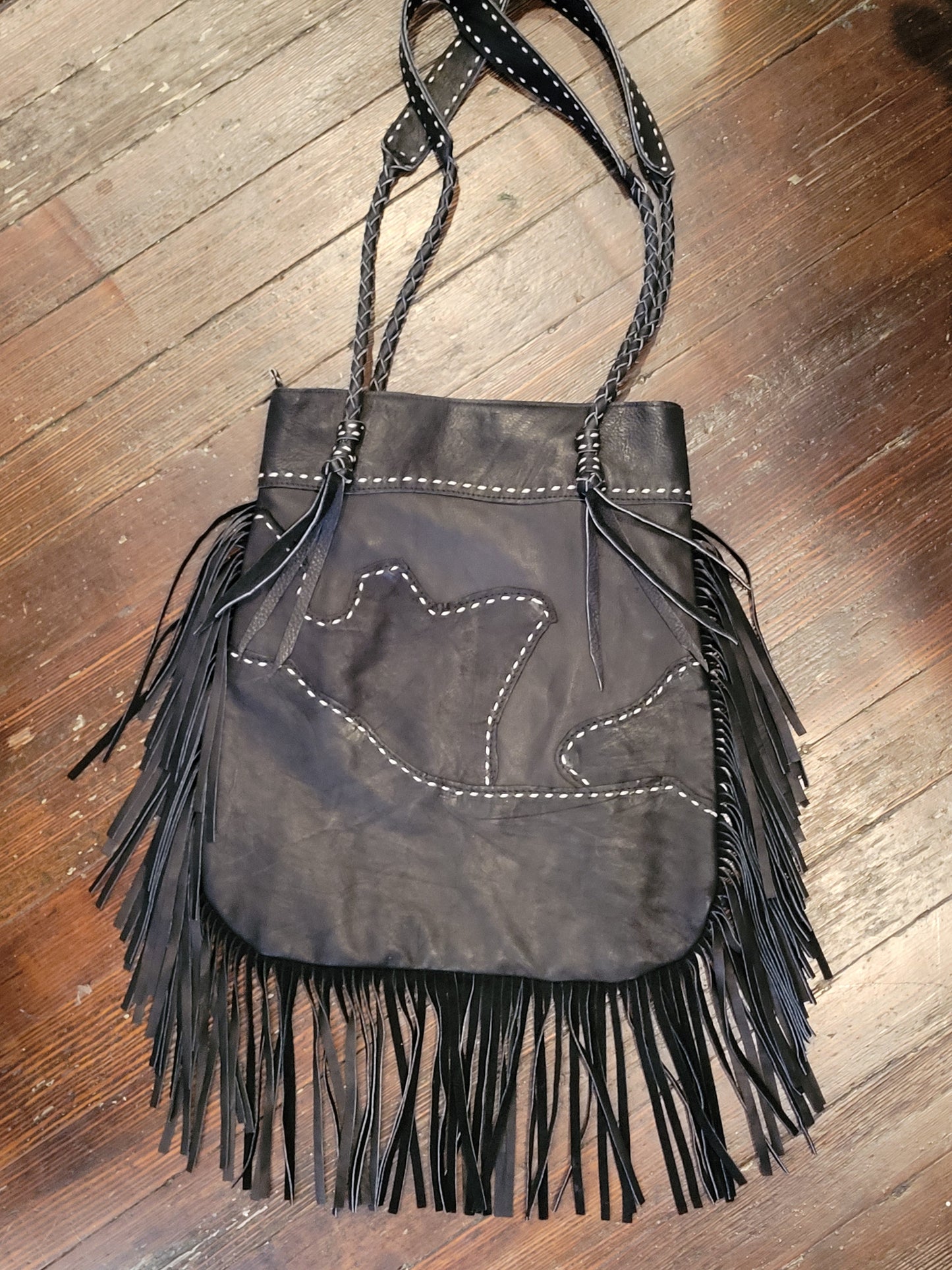 A black leather fringe handbag with white contrast stitching and braided shoulder straps, laid flat on a wooden floor.