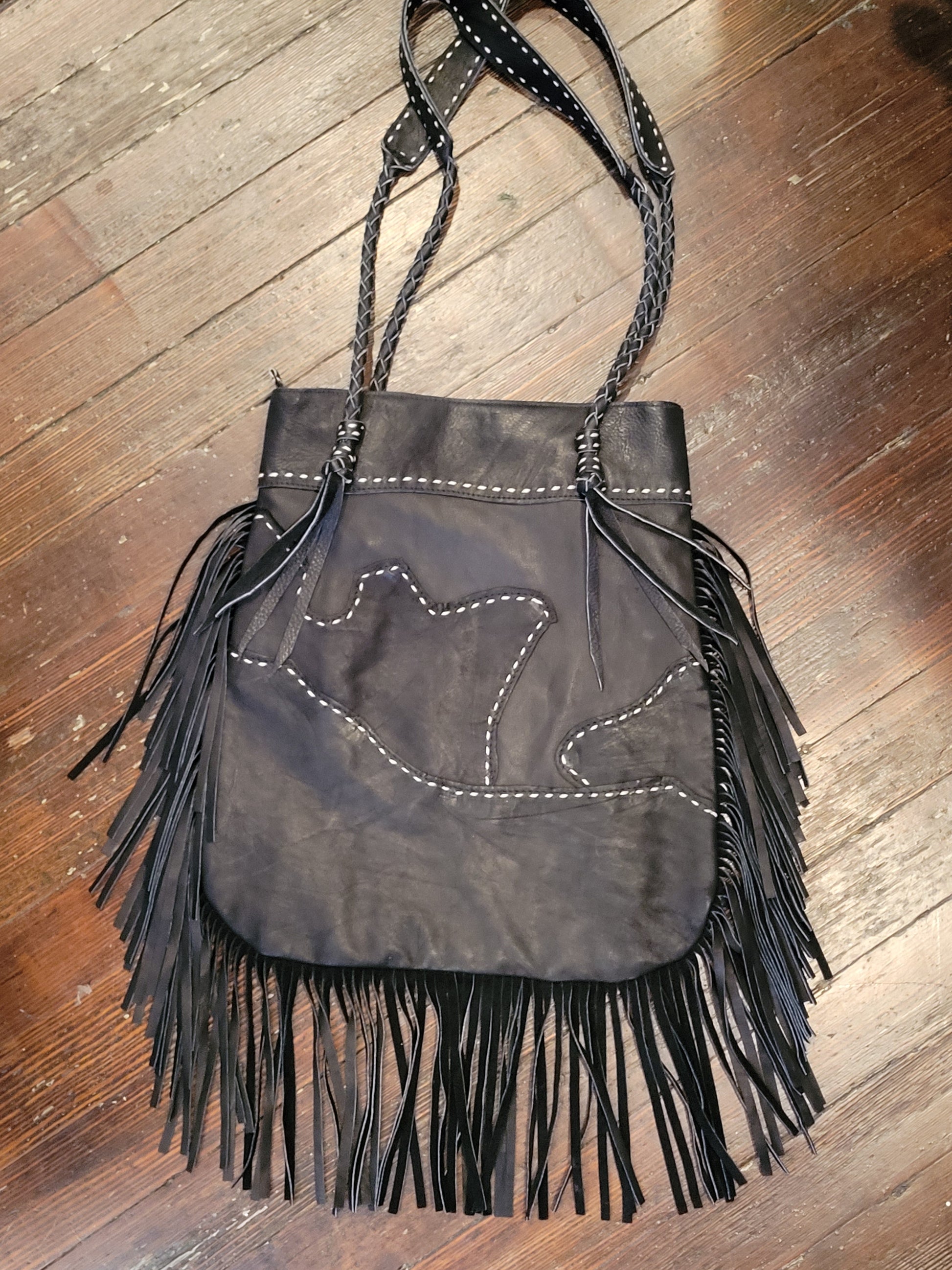 A black leather fringe handbag with white contrast stitching and braided shoulder straps, laid flat on a wooden floor.