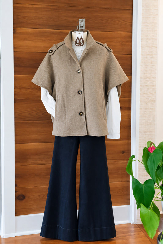 A Toffee short-sleeve button-front cape jacket with shoulder epaulets, styled over a white turtleneck and black wide-leg pants on a hanger, set against a wooden wall background.