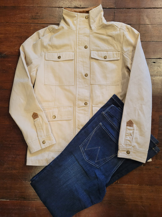 A cream-colored canvas Barn Jacket/Coat with a stand-up collar and button front, laid flat on a dark wood floor next to a pair of dark blue jeans.