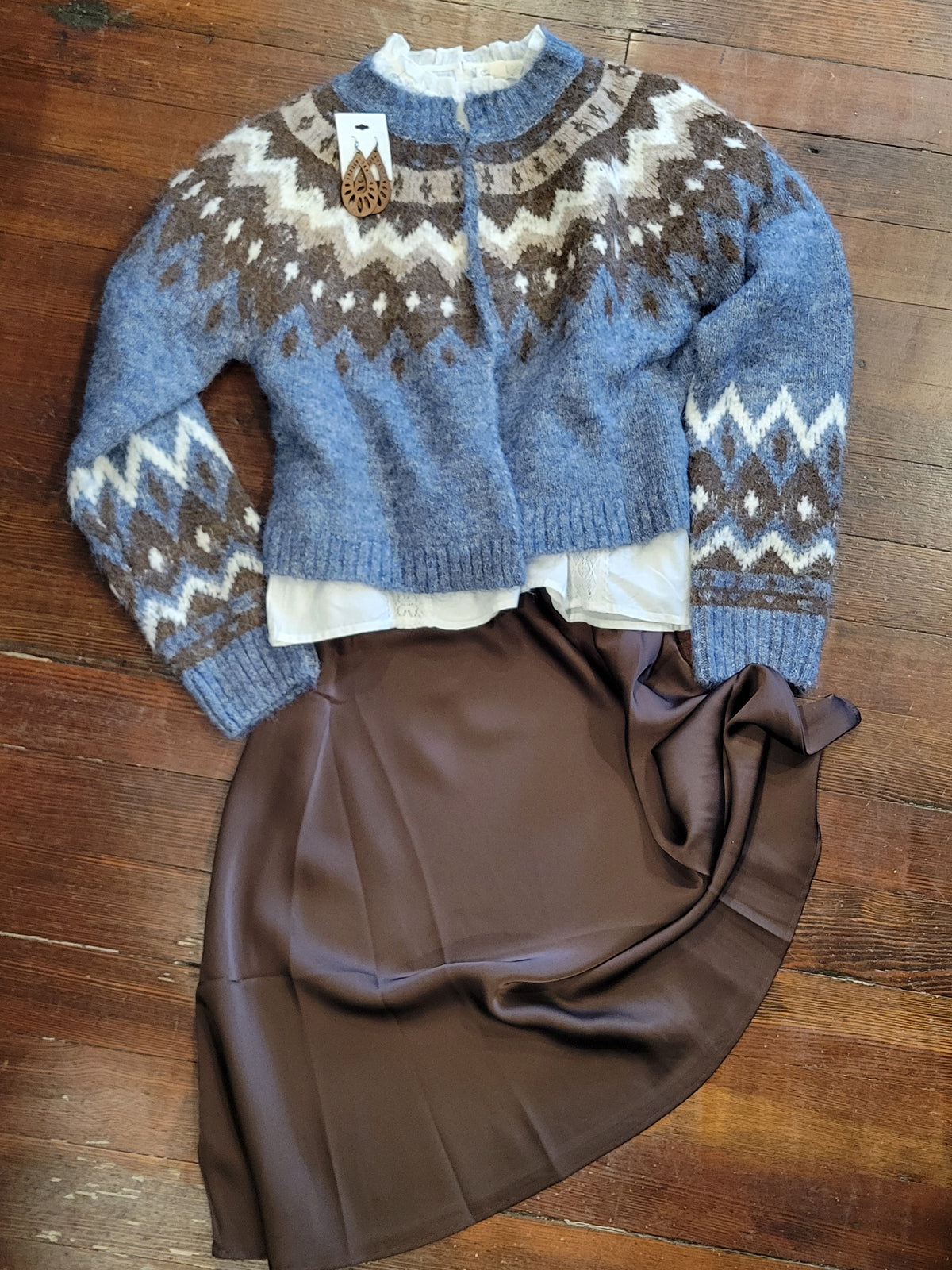 A blue knit cardigan sweater with brown and cream faire isle patterns, laid flat on a wooden floor next to a brown satin skirt.