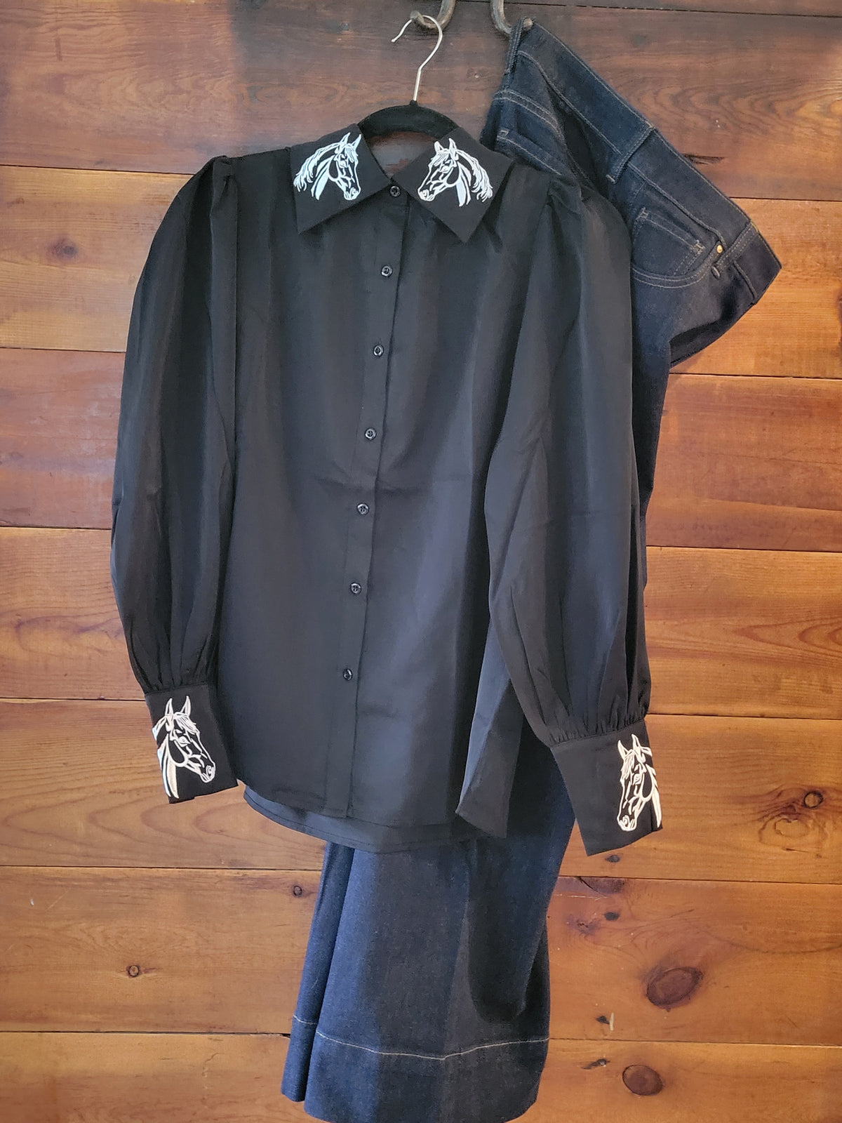 A black long-sleeve button-up shirt with white embroidered horse motifs on the collar and cuffs, displayed on a hanger against a wooden plank wall, with a pair of dark denim pants draped behind it.