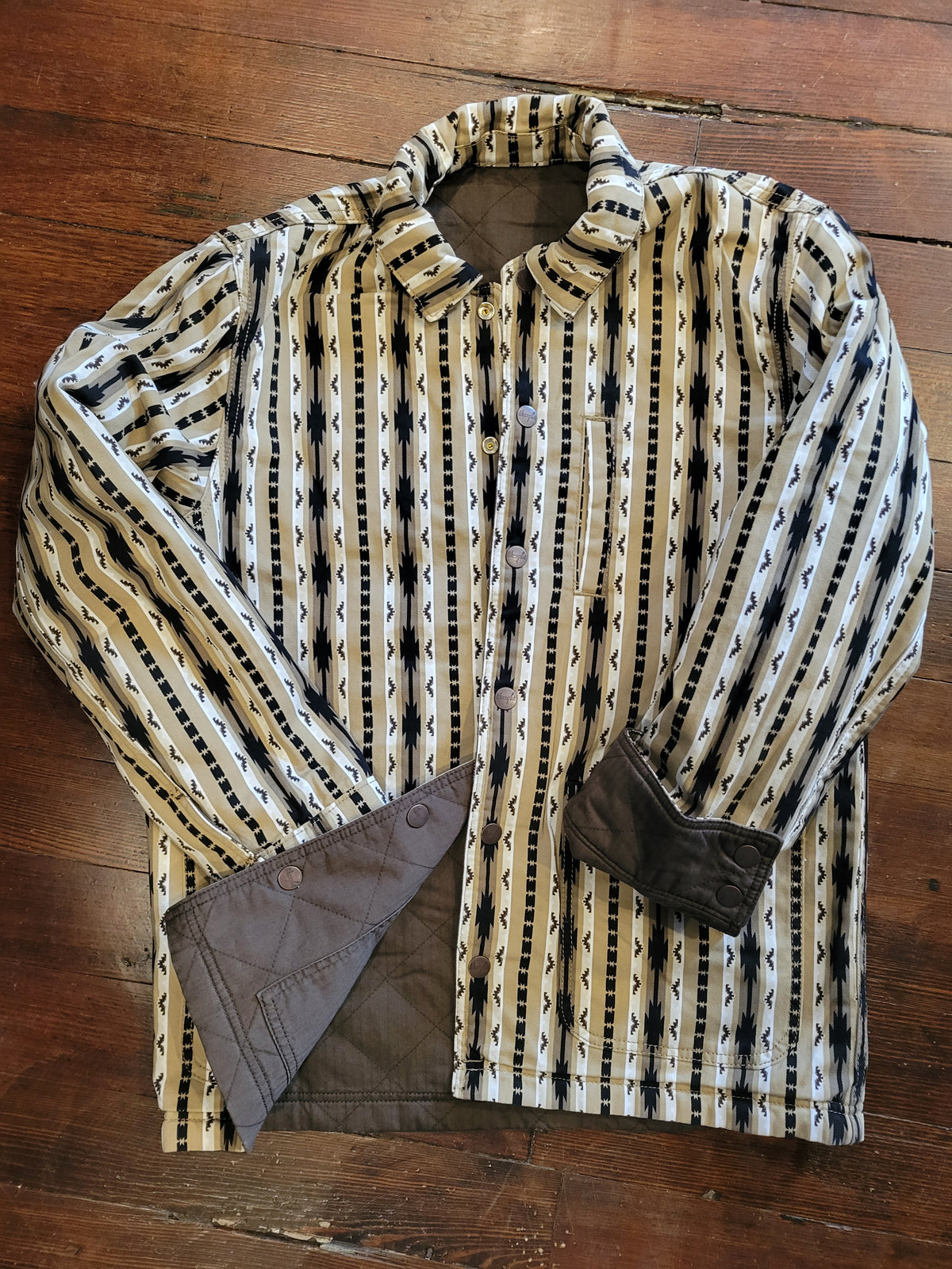 A long-sleeve button-up reversible jacket with a beige, black, and white geometric pattern, laid flat on a dark wood floor. The jacket is shown with the front flap turned up to reveal a solid dark brown lining on the reerse side.
