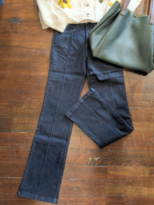 Dark wash blue jeans laid flat on a wooden floor, styled with a cream cardigan, a green leather tote bag, and a yellow flower.
