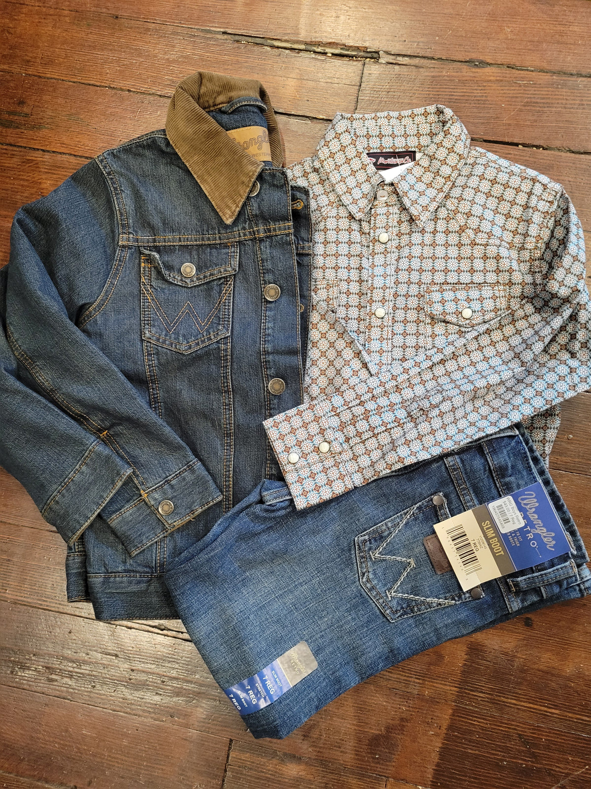 A flat-lay of two children's Wrangler outfits on a wooden floor, featuring a blue denim jacket with a brown corduroy collar, a light blue and white patterned long-sleeve shirt, and blue denim jeans with a blue brand tag still attached.