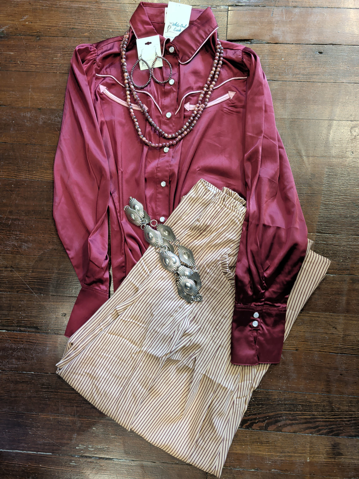 Tan pinstripe palazzo pants folded beneath a burgundy Western blouse, styled with a silver-tone concho belt, beaded necklace, and hoop earrings.