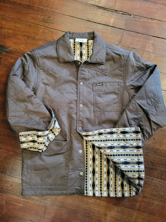 A brown quilted work jacket with a patterned lining, laid flat on a wooden floor. The jacket features a button-up front, chest pocket, and rolled-up sleeves showing the geometric pattern lining.