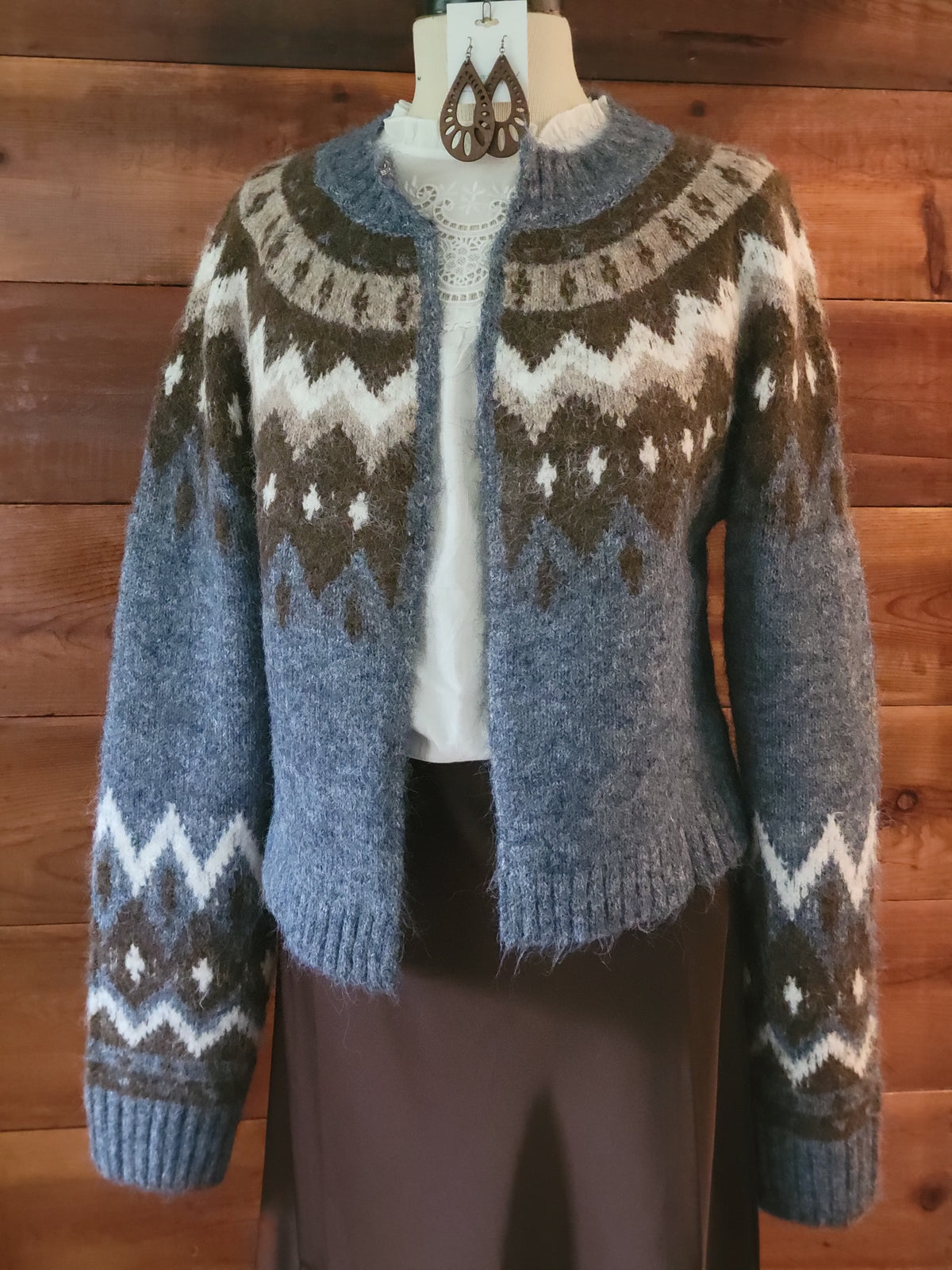 A fuzzy knit cardigan sweater with a brown, gray, and white Fair Isle pattern, displayed on a mannequin torso against a wooden wall background.