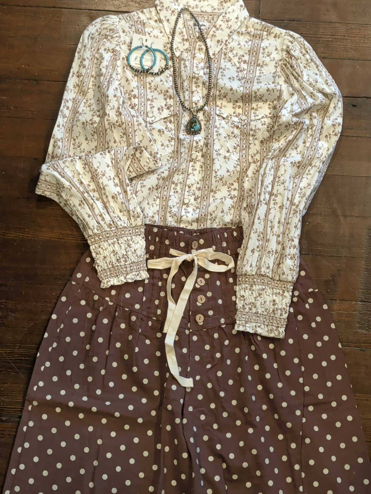 outfit close-up: Brown polka dot pants with a cream waist tie styled with a cream floral blouse and turquoise-colored jewelry.