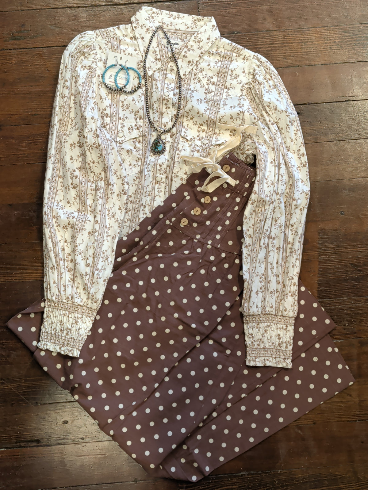Folded brown polka dot pants paired with a cream floral blouse and turquoise-colored jewelry on a wooden background.
