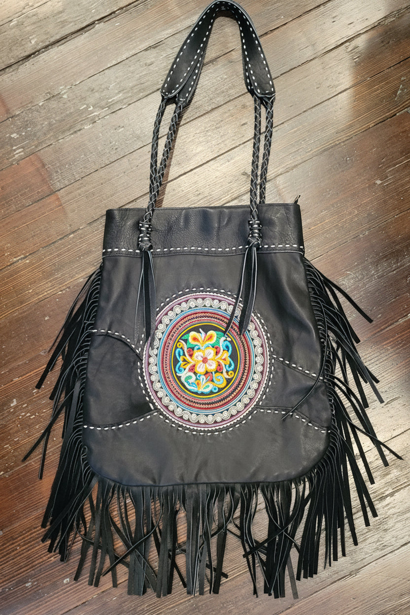 Black leather fringe handbag with a colorful circular embroidered patch on the front, resting on a wooden floor.